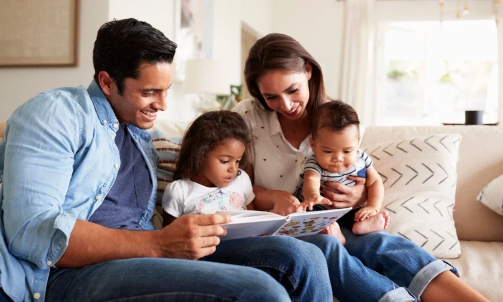 Young Hispanic family of four sitting on the sofa reading a book together in their living room