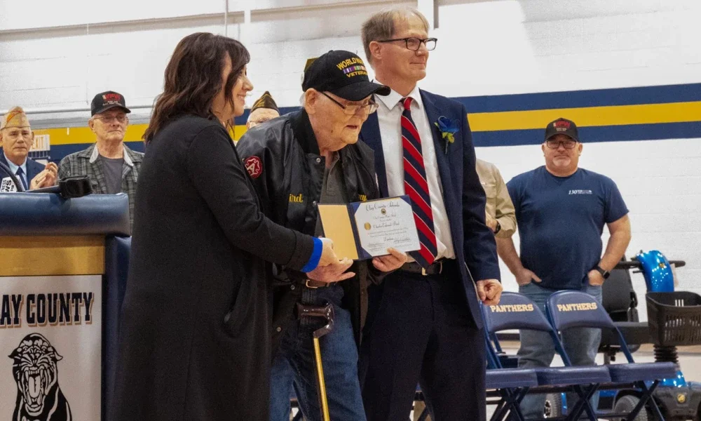 Older veteran receiving diploma at event.  Two people standing to either side to congratulate him, crowd behind