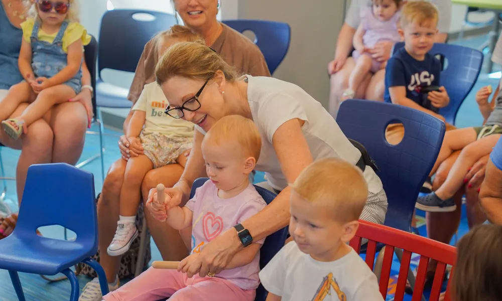 A women assisting a child with an activity. She is sitting in a chair surrounded by other adults and children sitting in chairs.