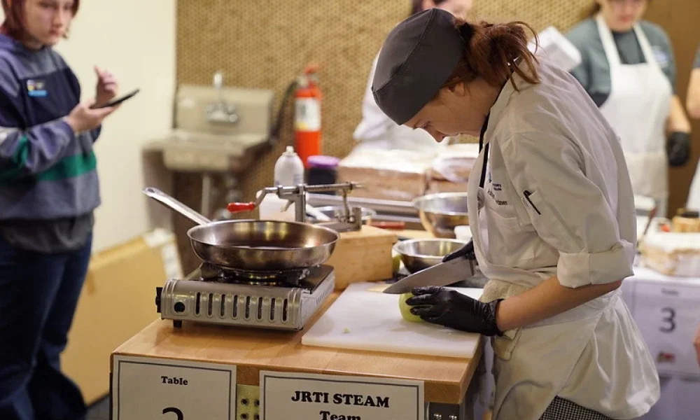 A student wearing a chef's outfit. She is wearing gloves while cutting an apple with a knife.