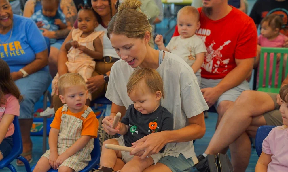 A mother and child playing with musical sticks while sitting on the ground. They are surrounded by other children and families.