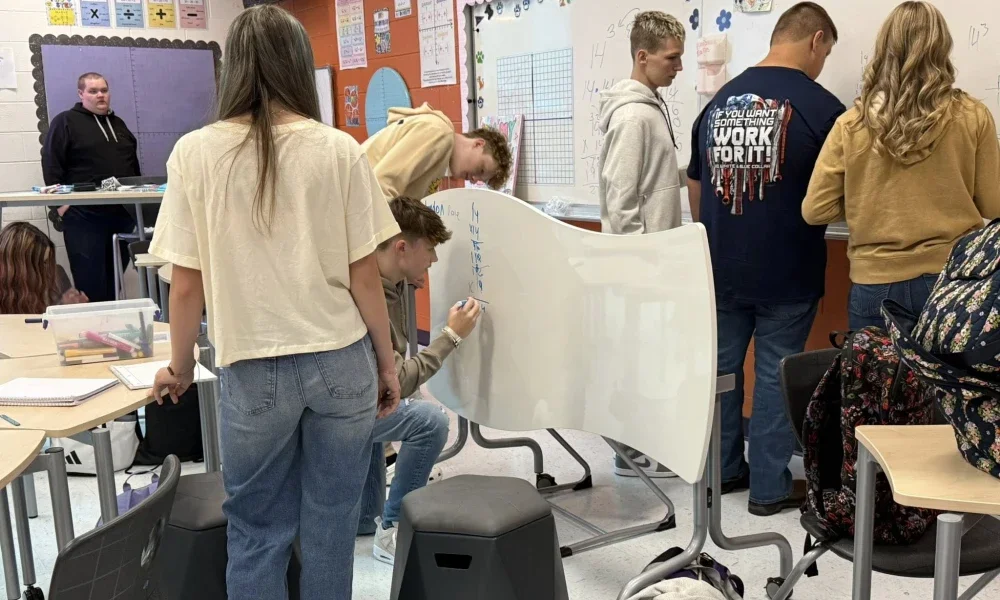 Students in a classroom solving math problems. One student is writing on a desk that doubles as a whiteboard.