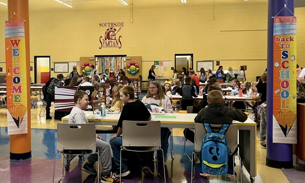 Students sitting at tables eating food inside of a school cafeteria.