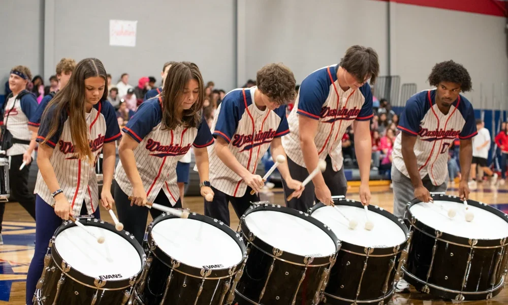 Students in a line playing bass drums hanging from a shoulder harness.