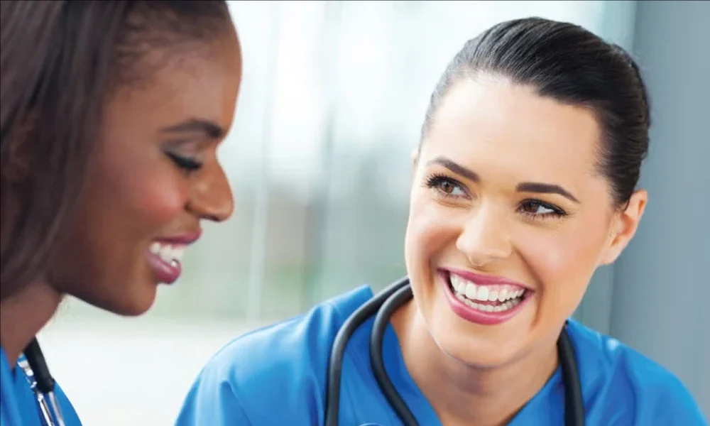 A nurse with a stethoscope smiles and directs the attention of another nurse towards a medical record.