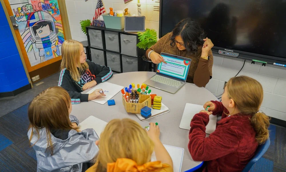 A teacher pointing at a screen while working with students who are sitting at a table.