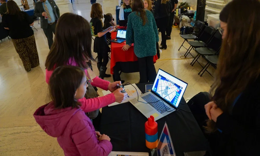 Two people standing in front of a computer while one person is holding a video game controller.