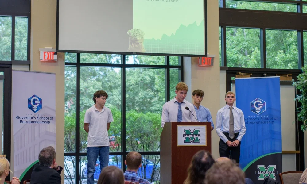 Three students on a stage with one student speaking from a podium.