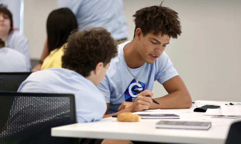 Students sitting at a table while one is pointing a pen at a piece of paper. 