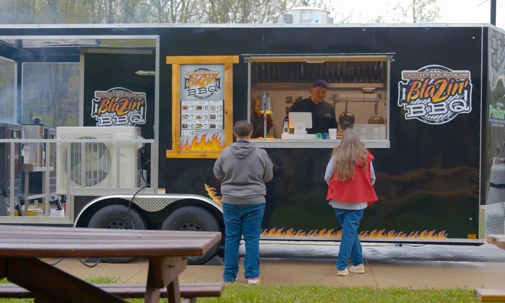 Black food trailer serving food words Blazin BBQ printed on side of trailer. Two customers wait to be served