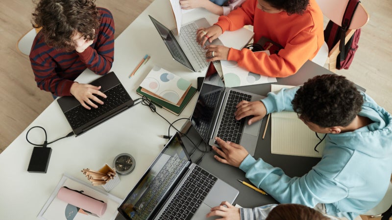 Top view at diverse group of teen school children using computers in classroom studying together at table for Governor's School of Computer Science.