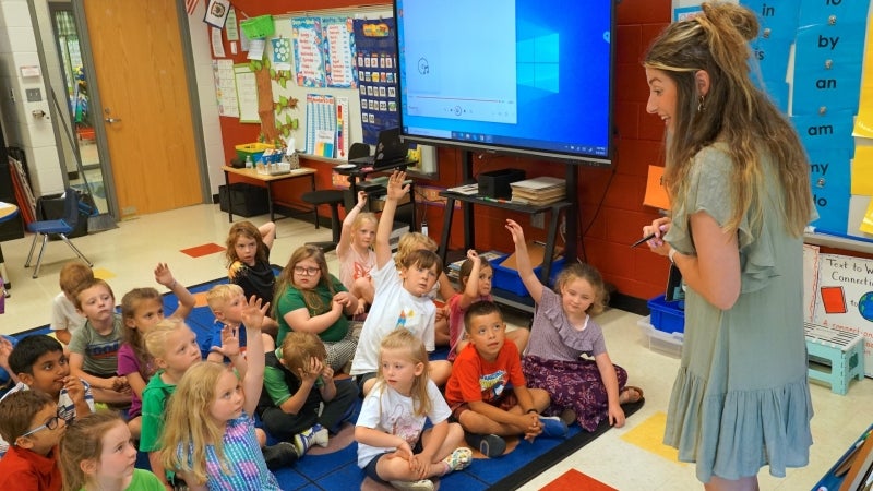 A teacher sharing a lesson with students. The teacher is standing at a whiteboard and the students are sitting on the classroom rug participating.