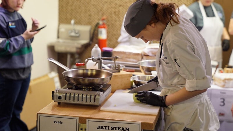 A student wearing a chef's outfit. She is wearing gloves while cutting an apple with a knife.