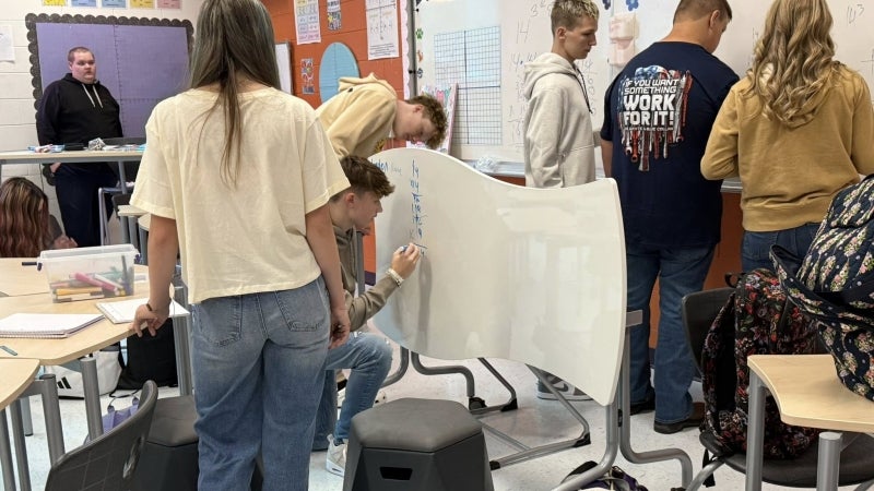 Students in a classroom solving math problems. One student is writing on a desk that doubles as a whiteboard.