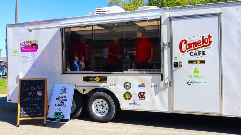 White food trailer.  Camelot Cafe is written on the side. Several students can be seen from the window getting ready to serve customers.