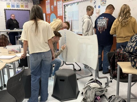 Students in a classroom solving math problems. One student is writing on a desk that doubles as a whiteboard.