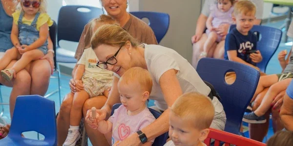 A women assisting a child with an activity. She is sitting in a chair surrounded by other adults and children sitting in chairs.