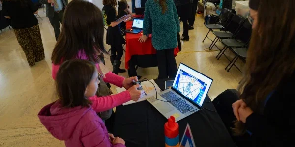Two people standing in front of a computer while one person is holding a video game controller.