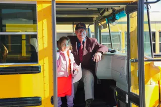 A student with a backpack stands inside a school bus beside an adult driver seated near the front