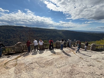 Students leaning against a fence while standing on a cliff looking at a mountain range.
