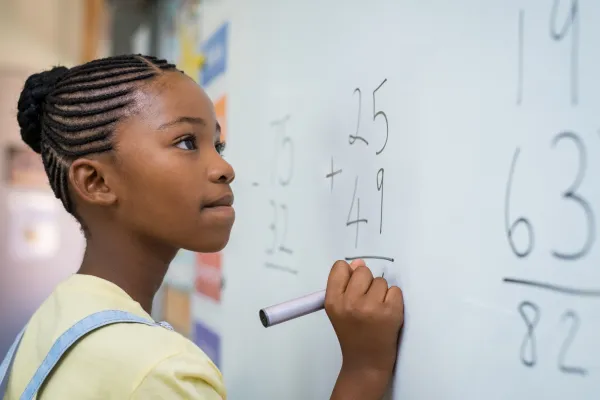 A young female holds a marker while staring with determination at an addition problem on the whiteboard.