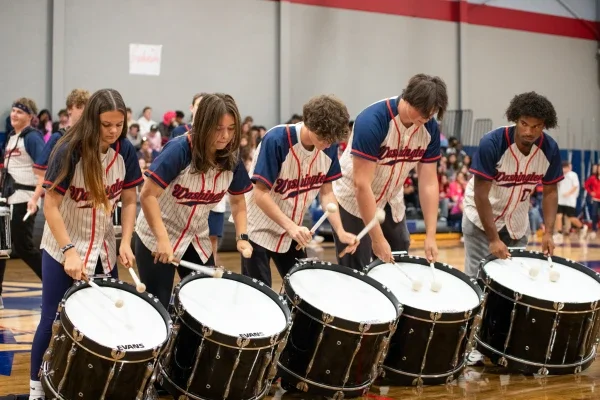 Students in a line playing bass drums hanging from a shoulder harness.