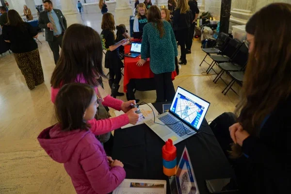 Two people standing in front of a computer while one person is holding a video game controller.