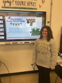 A student smiling in front of a TV screen. She was presenting about schools she plans to attend after high school.