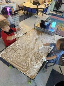 Students sitting at a table practicing writing. The table is covered in shaving cream.