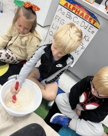 Students sitting on the floor while one student mixes dry ingredients in a bowl.