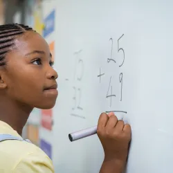 A young female holds a marker while staring with determination at an addition problem on the whiteboard.