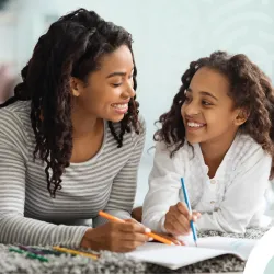 Mother and child glance at one another while working on a document together and lying on the floor.