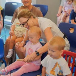 A women assisting a child with an activity. She is sitting in a chair surrounded by other adults and children sitting in chairs.