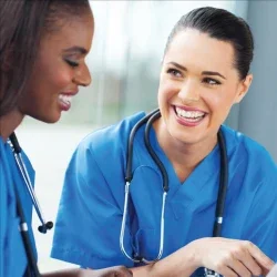 A nurse with a stethoscope smiles and directs the attention of another nurse towards a medical record.