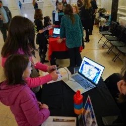 Two people standing in front of a computer while one person is holding a video game controller.